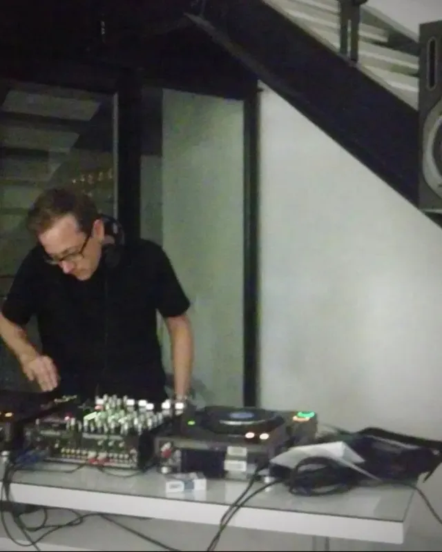 Andy, a.k.a. DJ Dusty, leaning over a mixer and CDJs at night beneath a staircase, a studio monitor on a stand beside him